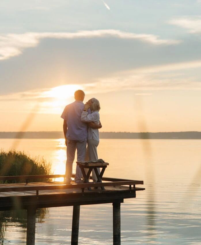 An older couple embracing on a lakeside jetty at sunset.